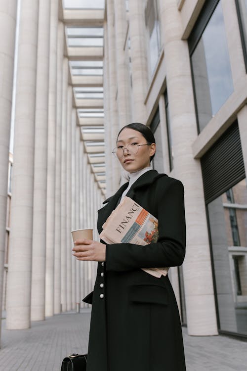 A Low Angle Shot of a Woman in Black Coat Holding a Cup of Coffee while Standing Near the Building