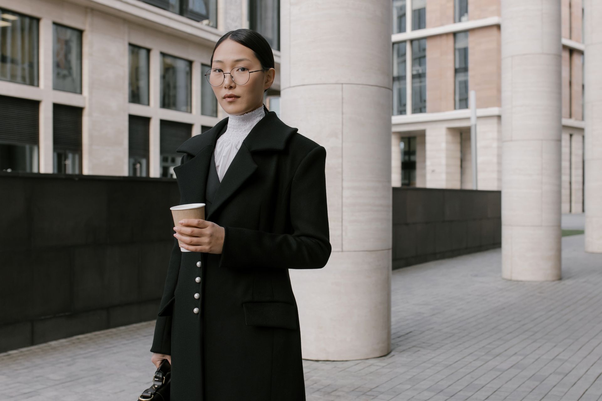 Asian businesswoman holding a coffee cup and standing outdoors in a modern urban area.