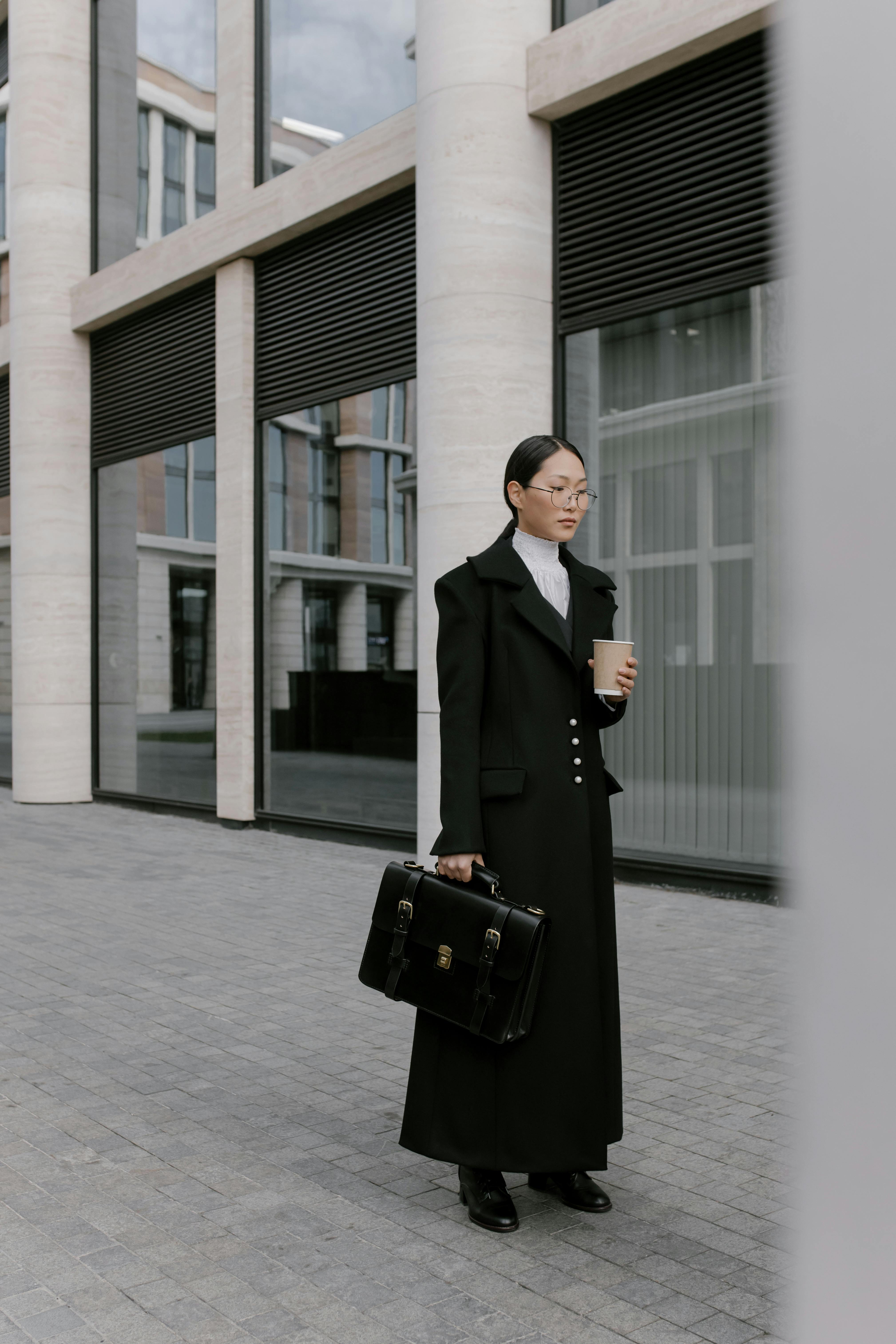 woman in black coat with briefcase holding coffee cup and standing beside building with big windows