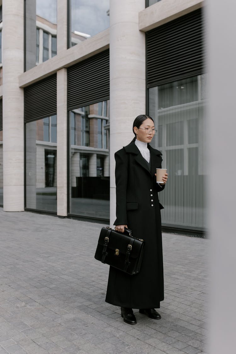 Woman In Black Coat With Briefcase Holding Coffee Cup And Standing Beside Building With Big Windows