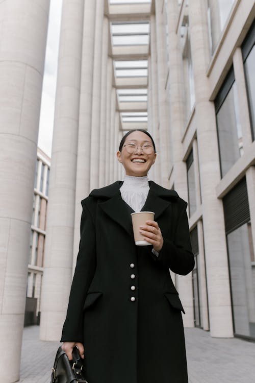 Free A Low Angle Shot of a Happy Woman Standing Beside the Building while Holding a Cup of Coffee Stock Photo