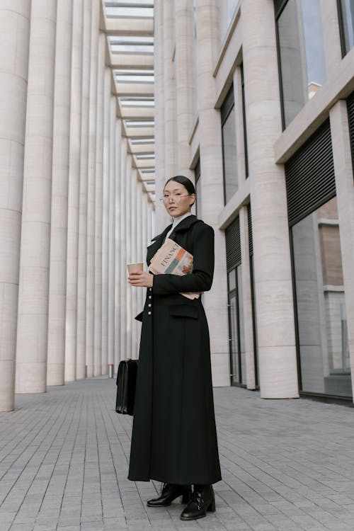 Woman in Black Coat Holding Newspaper and Coffee to Go