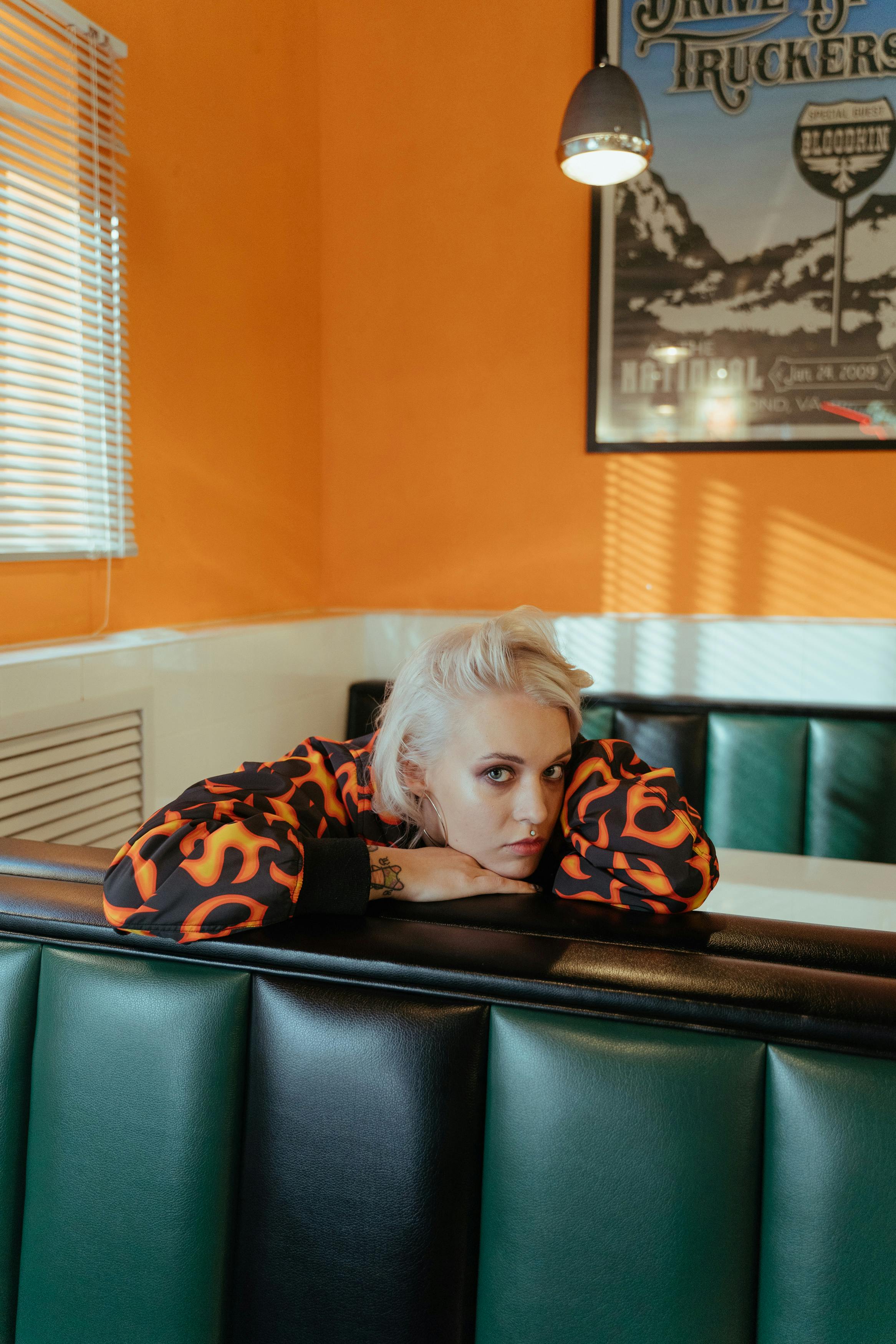 Blonde woman in a vibrant diner booth, wearing stylish orange and black outfit, with a contemplative expression.