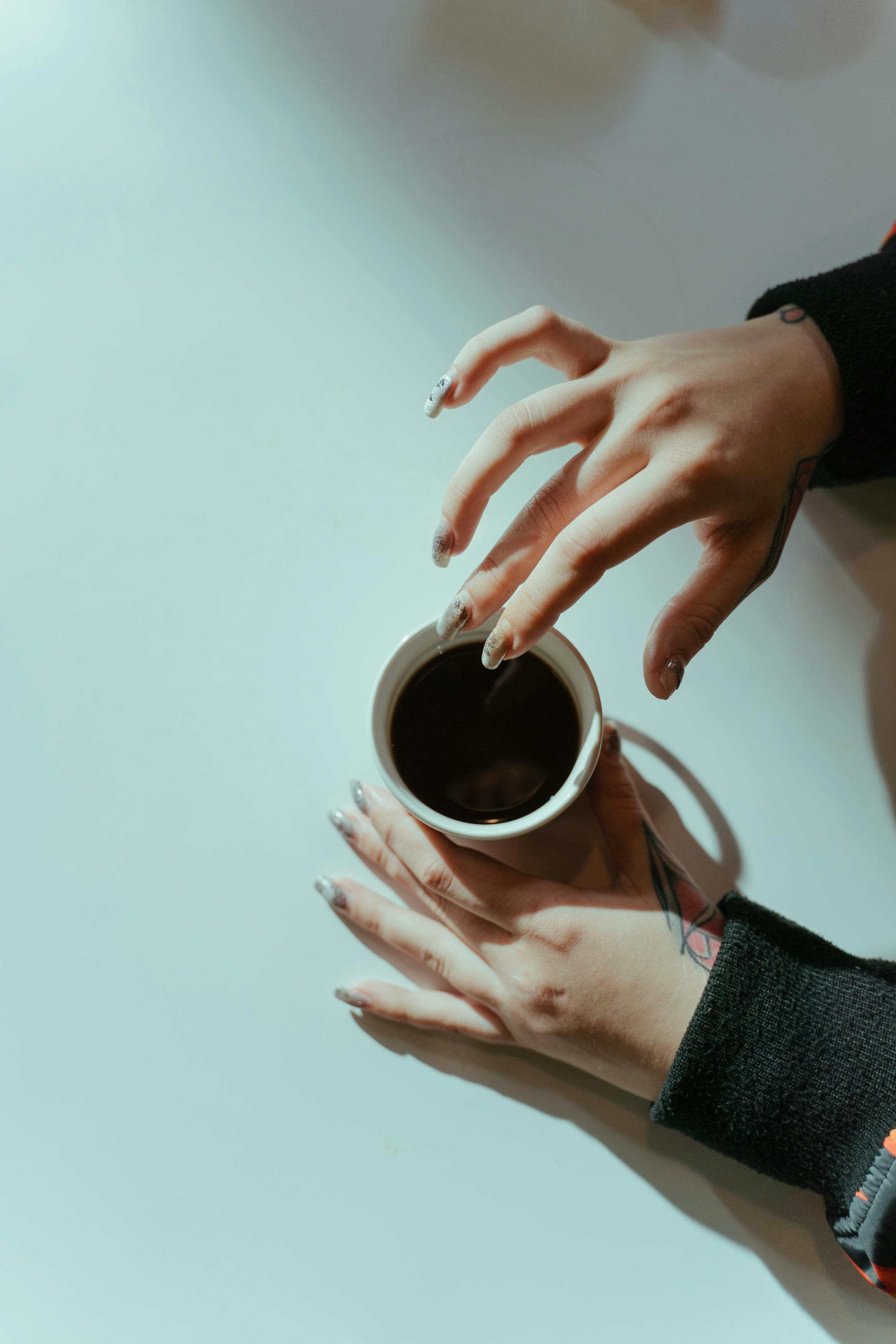 Hands with tattoos holding a coffee cup on a white surface, captured from above.