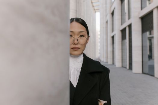 Elegant woman in black coat and eyeglasses stands in modern urban architecture.