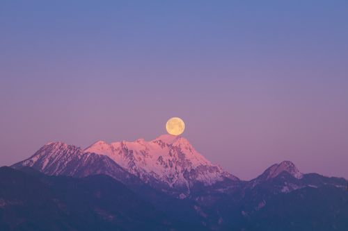 Free A stunning full moon rises above snow-covered mountain peaks during twilight, casting a serene pink hue. Stock Photo