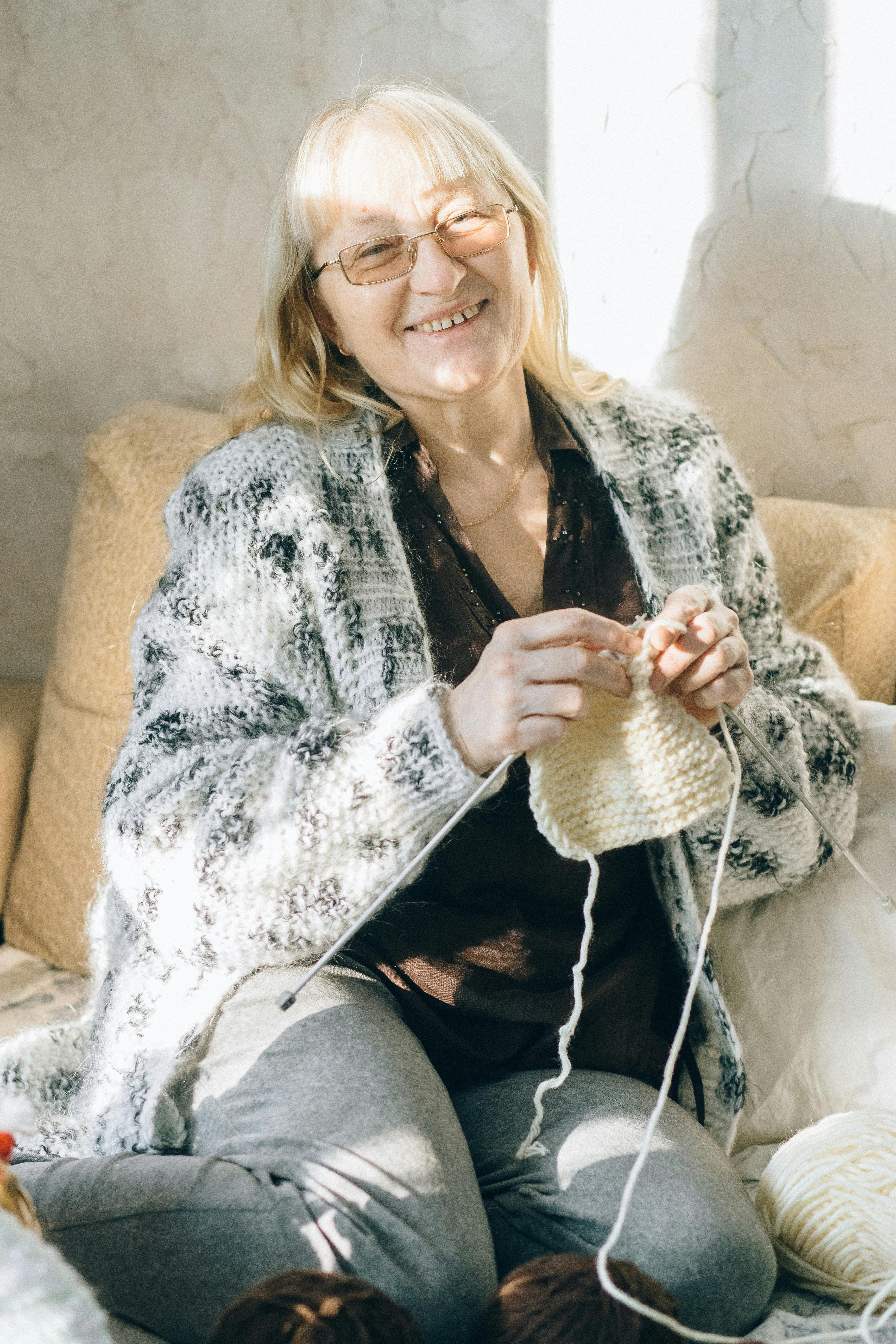 elderly woman in white and black cardigan sitting on a couch doing crochet