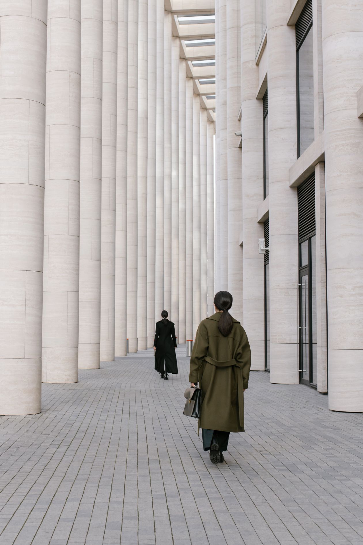 Two women in trench coats walking on a city street lined with architectural columns
