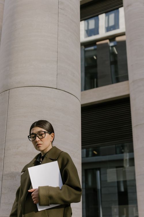 Free Asian woman with glasses and coat standing outside among modern columns holding a white folder. Stock Photo