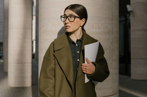 Free A professional woman in a coat standing outside, holding papers and looking away thoughtfully. Stock Photo