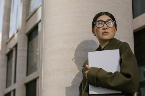 Free Asian woman in coat and eyeglasses holding documents outdoors, looking afar contemplatively. Stock Photo