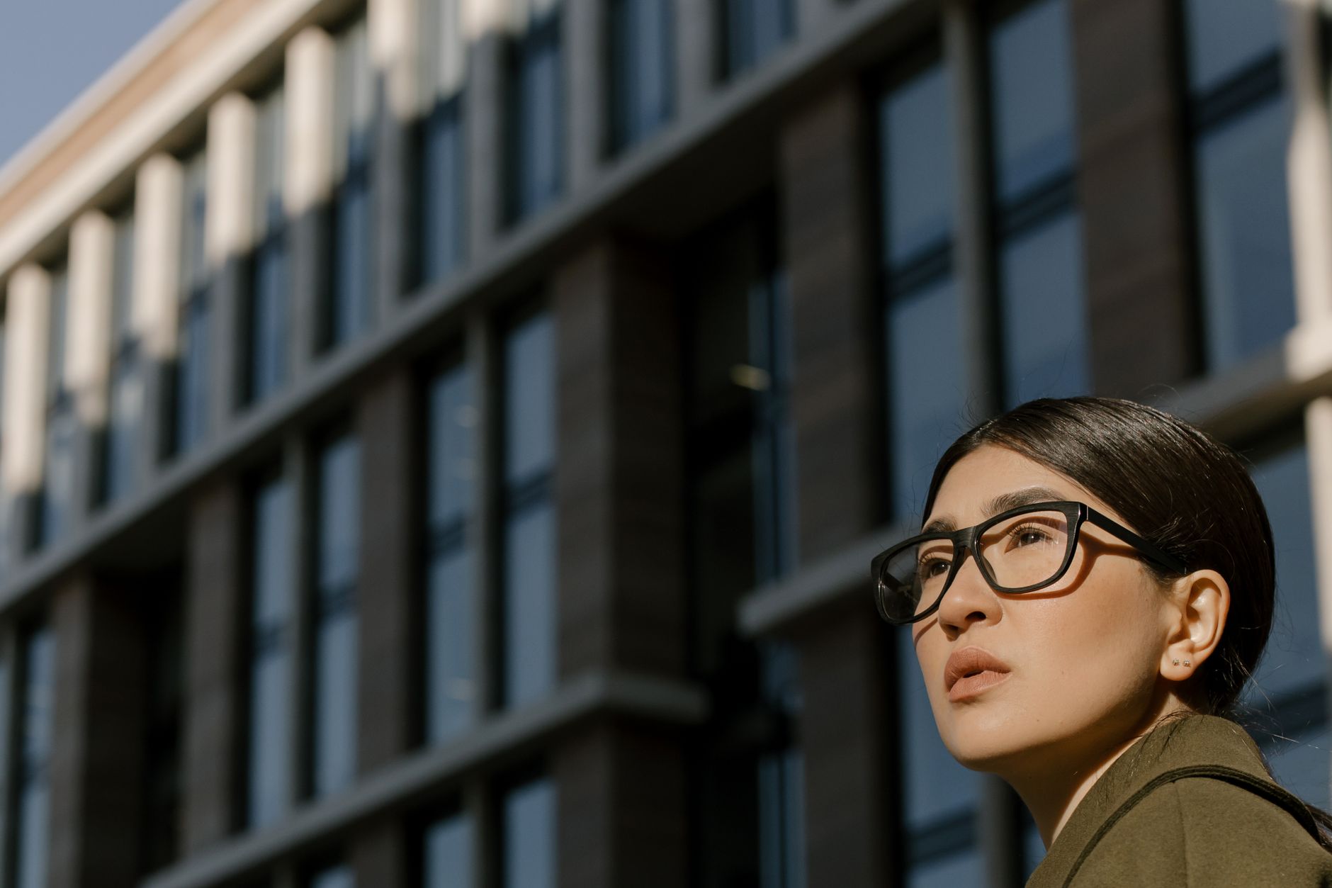 Low-angle shot of a serious woman in eyeglasses against a modern building.