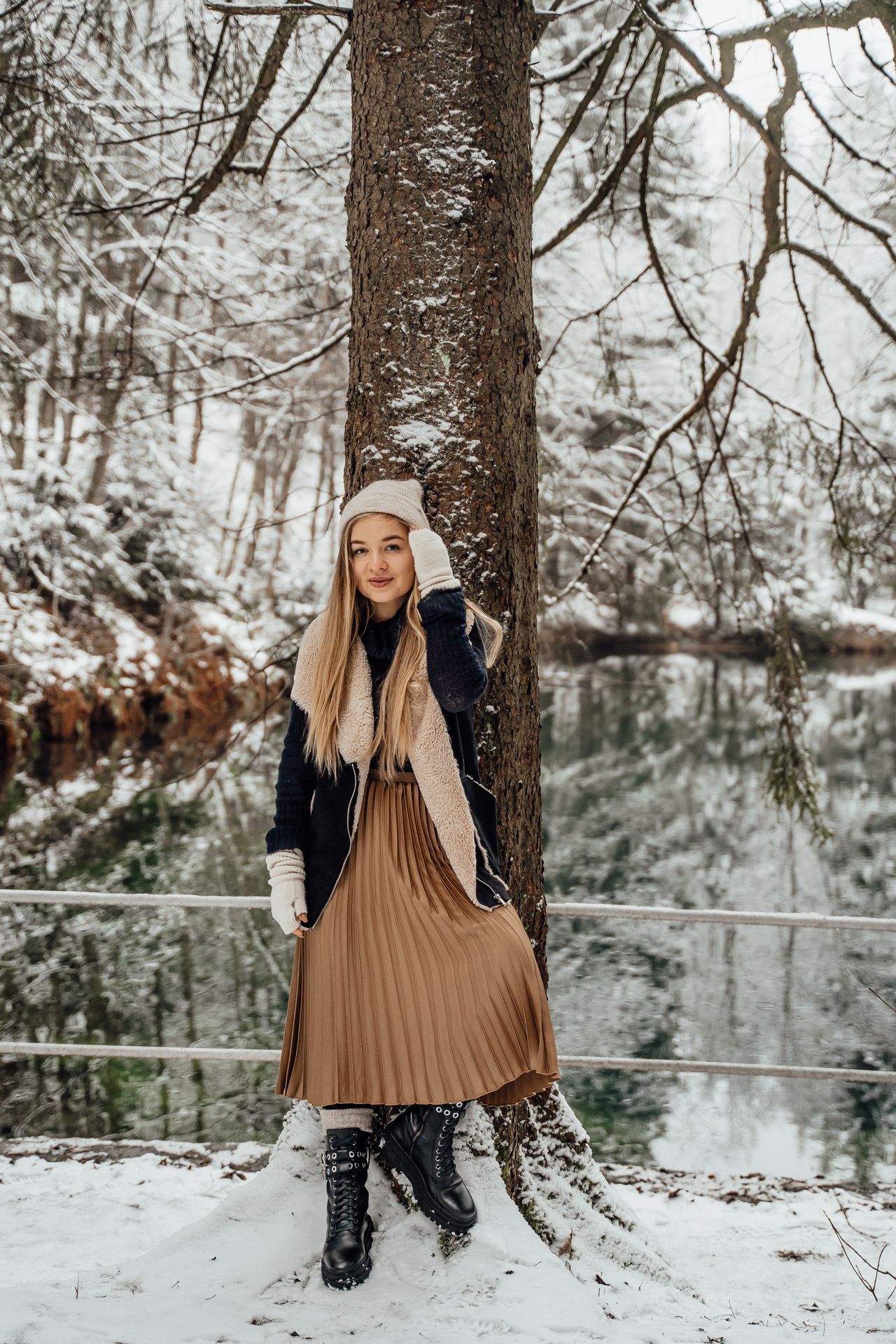 Woman in snow wearing a camel pleated midi skirt, navy sweater, shearling vest and boots
