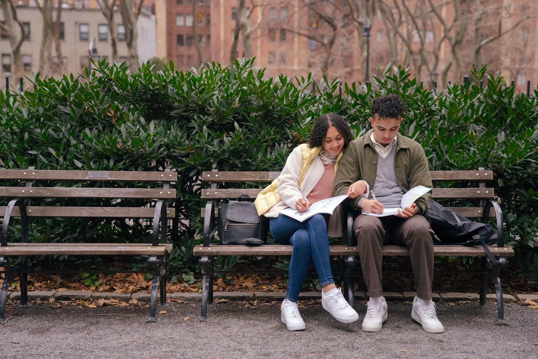 Student couple preparing homework on bench in city park