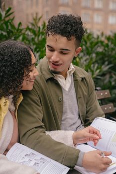 Smiling ethnic couple of students in casual wear sitting on bench and writing in workbooks while working on project in city park