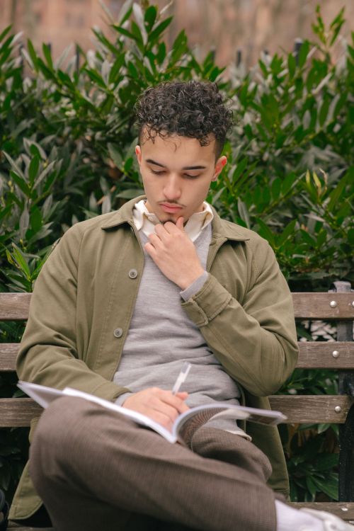Free Thoughtful ethnic curly student male in casual outfit sitting on bench and reading textbook while preparing homework in city park at daytime Stock Photo