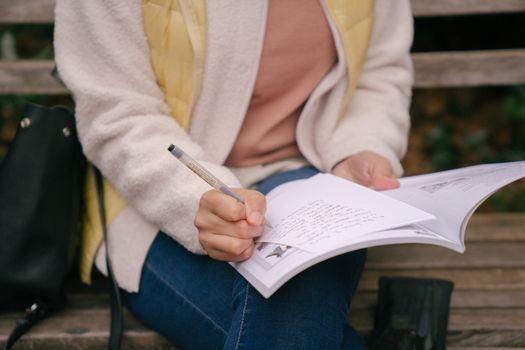 High angle of crop unrecognizable concentrated woman in casual outfit writing in workbook while preparing homework on wooden bench in park at daytime