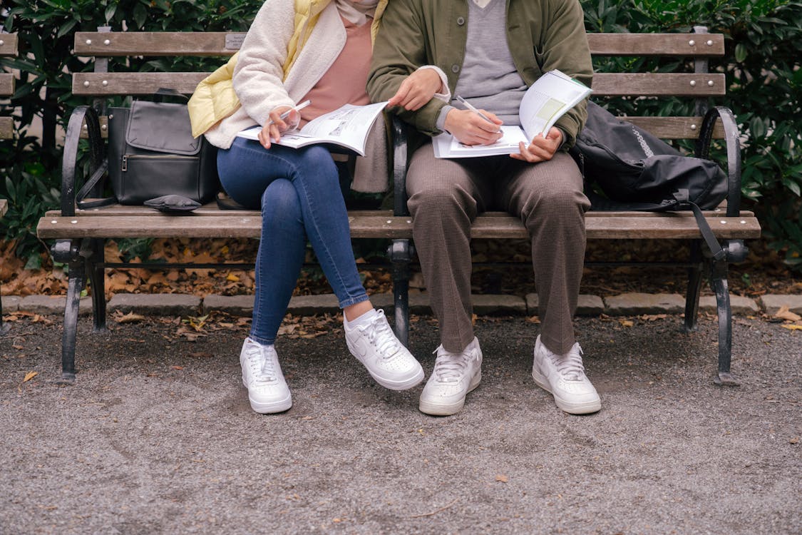 Free Crop couple in casual clothes and white sneakers sitting on bench in city park while doing homework in workbooks at daytime Stock Photo