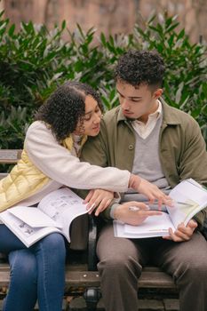 Happy ethnic student couple in casual outfit preparing homework together while sitting on bench in park and checking exercises in workbooks