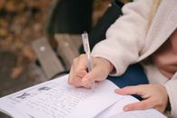 Crop unrecognizable woman in casual outfit sitting on wooden bench while taking notes on paper near textbook in street with foliage on ground in daylight in autumn