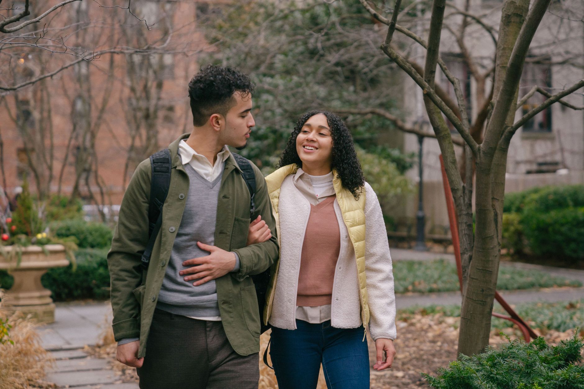 Young Hispanic couple in casual outfit walking in park on walkway while having date and holding hands and looking at each other near bushes and tree in daylight near buildings