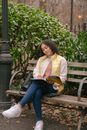 Full body of dreamy young Hispanic woman in casual outfit sitting on wooden bench while reading book near purse in park with foliage near green bushes and pole in daytime