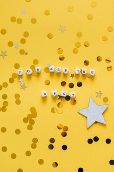 Flat lay of 'You Are The Best' cubes with stars on yellow surface. Motivational decor.