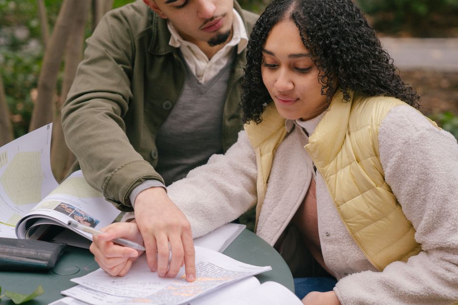 Young Latin American couple in casual outfit studying at table while reading papers and textbooks on street near bushes and trees