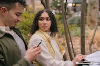 Side view of Hispanic female sitting at table with textbooks while preparing for exams with phone while looking jealously at man texting on cellphone in daytime in park