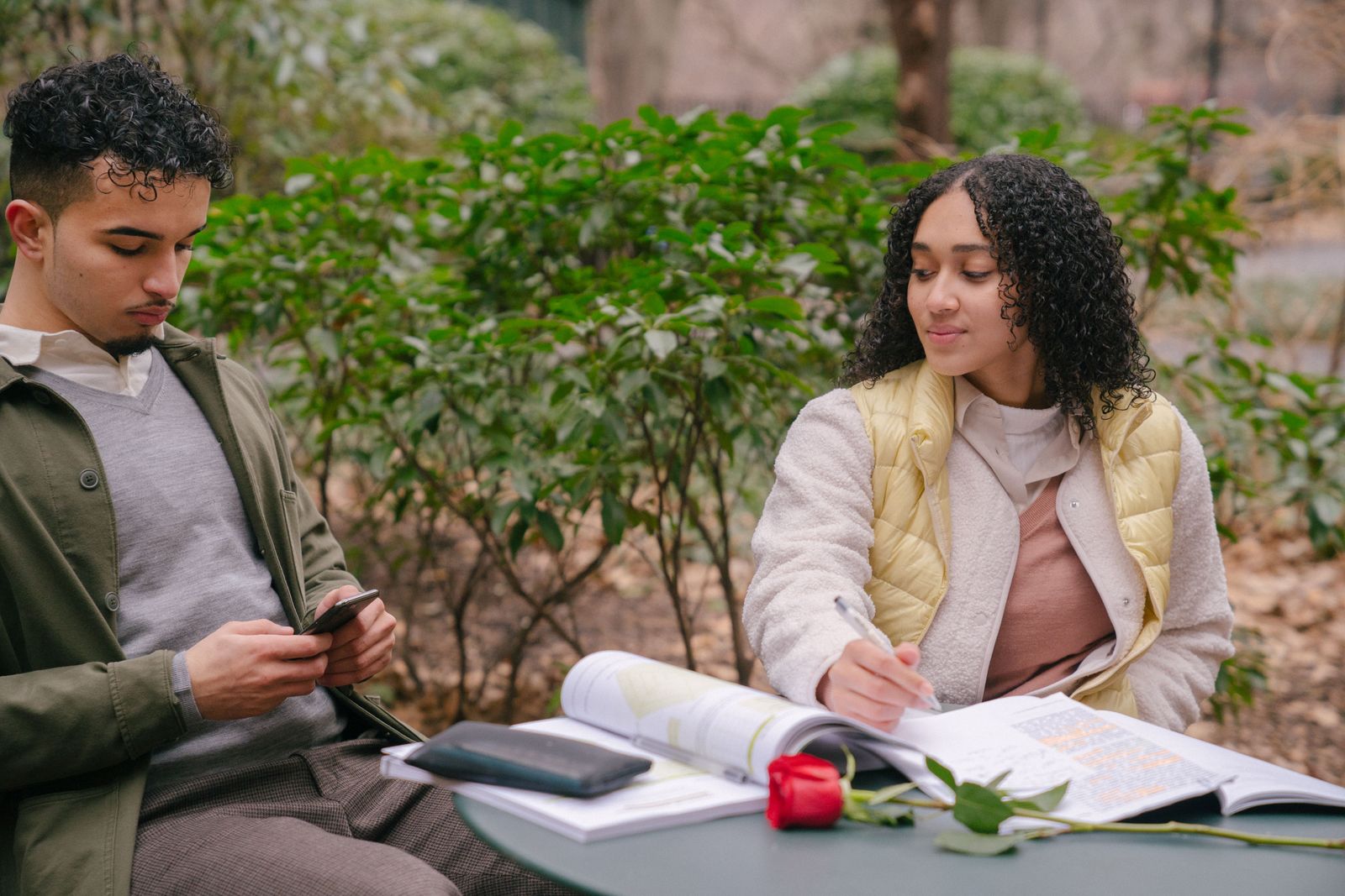 Two people using a phone and writing in a notebook at an outdoor table