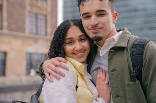 Free Cheerful Hispanic couple in casual clothes embracing each other while standing on city street and looking at camera in daylight Stock Photo