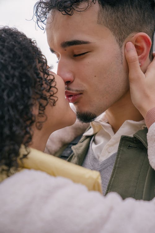 Cheerful young Hispanic couple in casual clothes embracing and kissing each other against light background in daytime