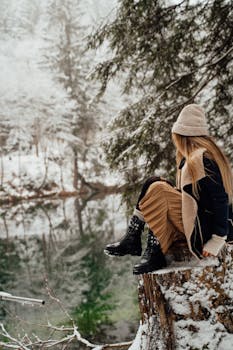 A woman sitting on a tree stump by a serene winter lake in a snowy forest.