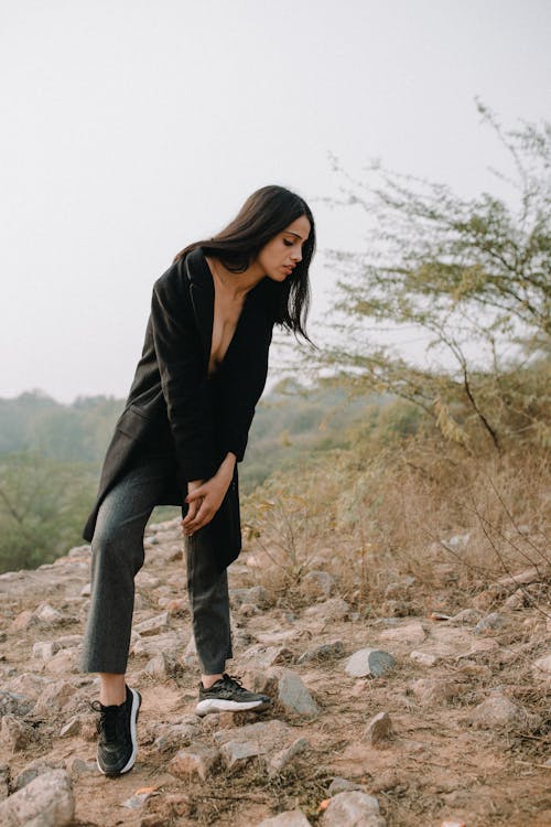 Full length of young serious female in black coat and pants standing on stony mountain near plants and grass in daylight in nature under gray cloudless sky