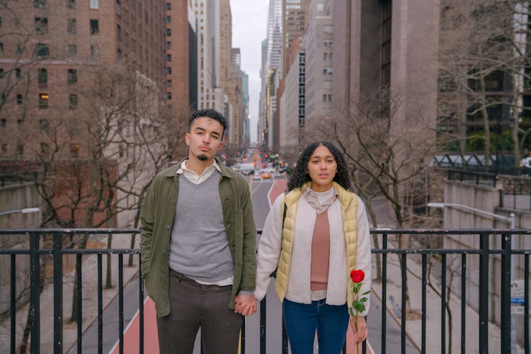 Pensive Hispanic Couple Holding Hands And Standing On Bridge Against City Street