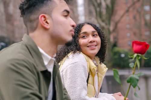 Free Side view of happy romantic ethnic lady in stylish warm clothes with red rose in hand smiling and looking at boyfriend during date in city park Stock Photo