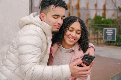 Free Joyful young Hispanic male and female millennials in stylish jackets smiling and embracing while watching video on mobile phone on street Stock Photo