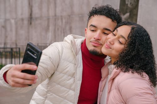 Free Enamored young Hispanic couple in warm casual clothes smiling and touching cheeks while taking selfie on mobile phone standing on street Stock Photo