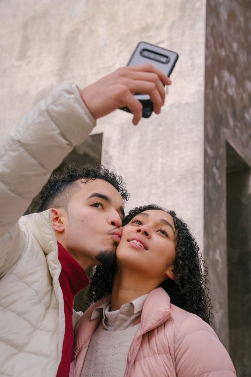 Free From below of enamored young Hispanic guy in casual clothes kissing cheek of happy girlfriend while taking selfie on smartphone on city street Stock Photo