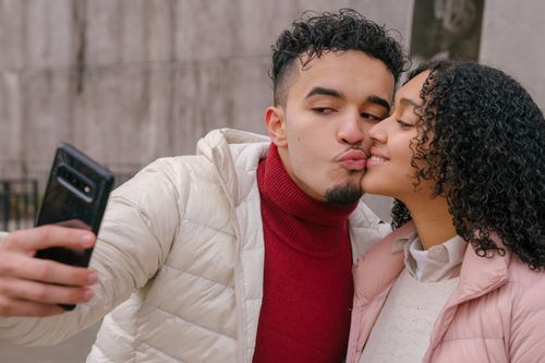 Free Happy young Hispanic couple in stylish warm jackets kissing and cuddling while chilling on street and taking selfie on mobile phone Stock Photo