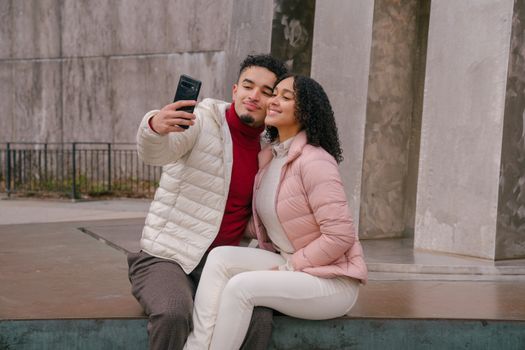Delighted young ethnic couple touching cheeks while taking selfie sitting on parapet