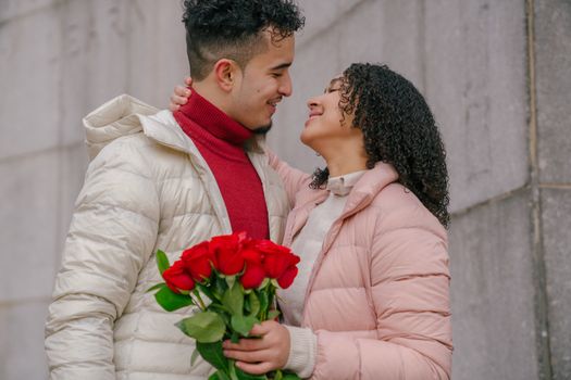 Side view of enamored young ethnic woman in warm clothes smiling and embracing happy boyfriend with presented bouquet of fresh red roses in hand
