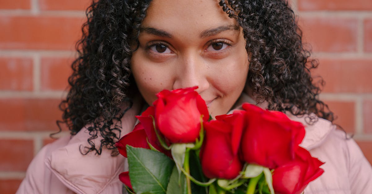 A young woman with curly hair, smiling and holding a bouquet of red roses in front of a brick wall.