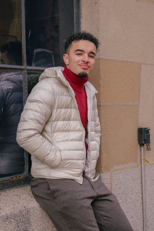 Confident young bearded ethnic guy in stylish warm clothes leaning on building with hands in jacket pockets and looking at camera