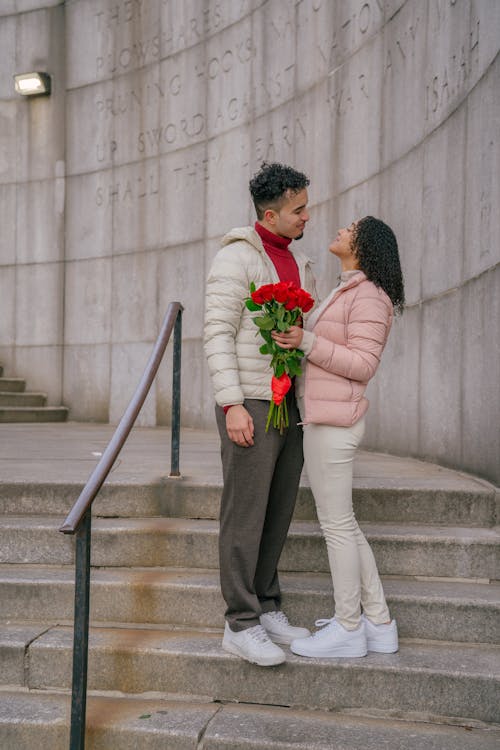 Side view of content young Hispanic woman with elegant bouquet of fresh red roses standing on stairs with romantic boyfriend during date on city street