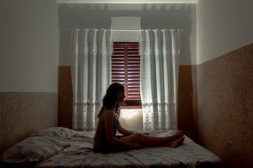 Free A woman sits on a bed, silhouetted by a window with curtains casting soft shadows. Stock Photo