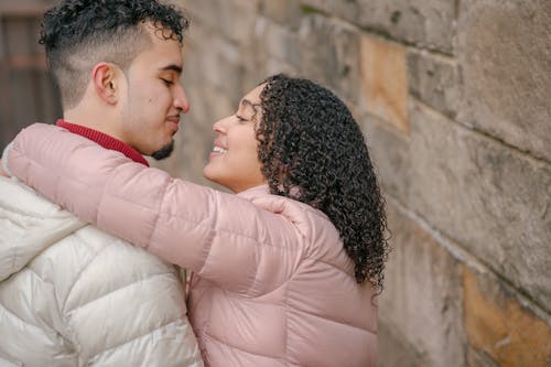 Side view of positive Hispanic couple wearing outerwear looking at each other and hugging while standing near stone wall on blurred background during romantic date