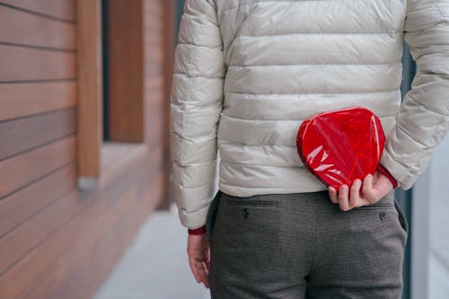 Free Back view of anonymous male hiding wrapped hear shaped present box while standing on street during holiday celebration in city Stock Photo