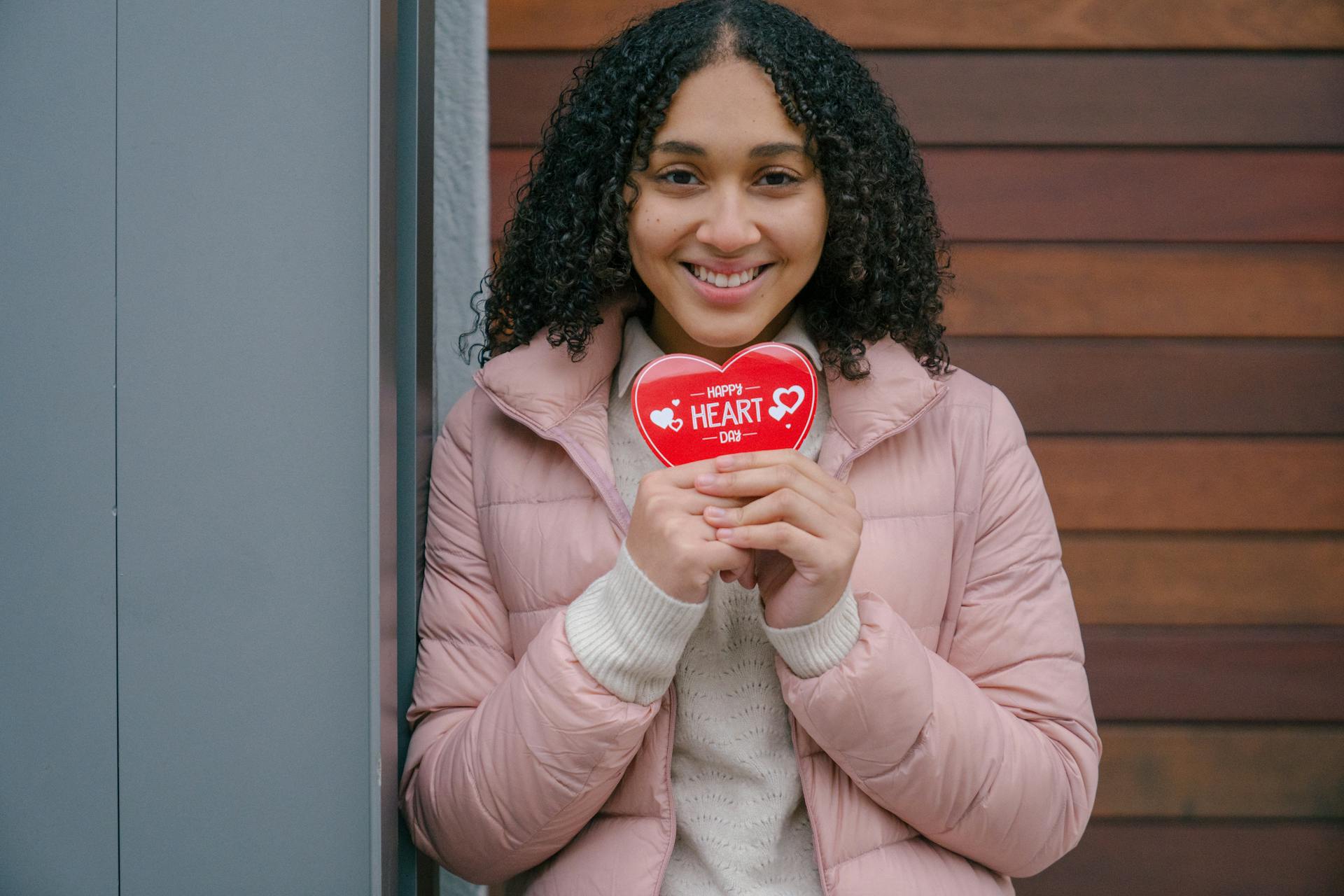 Cheerful Hispanic female wearing warm clothes looking at camera while standing on street with red heart shaped present in hands during Saint Valentine day