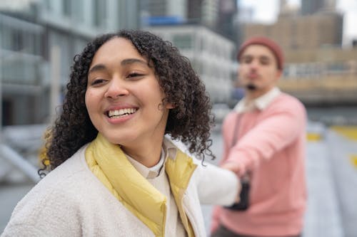 Free Optimistic Hispanic female with curly hair looking away while holding hands with unrecognizable boyfriend during romantic date on blurred background Stock Photo